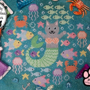 Puede incluir: Un patrón de punto de cruz con una sirena gato rodeada de criaturas marinas. El diseño incluye peces, cangrejos, medusas y estrellas de mar en tonos de rosa, amarillo y naranja sobre un fondo turquesa. El diseño incluye el texto "Simply Meowvelous Designs".