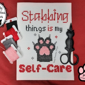May include: A cross-stitch pattern on white fabric featuring a black and pink cat paw print with claws and the text Stabbing things is my SELF-CARE. Included are four spools of embroidery thread in black, red, gray, and pink, and black cat-shaped scissors.