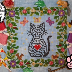May include: A cross-stitch pattern on light blue fabric featuring a black maze-like cat silhouette with a red heart in the center. The design is framed by a floral border with green leaves, purple and pink flowers, and butterflies.