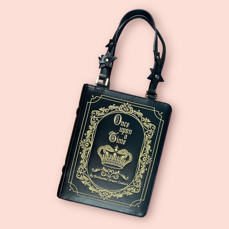 May include: Black book-shaped handbag with a gold chain strap and a gold inscription that reads "Once upon a Time by Enchanted Dream Couture".