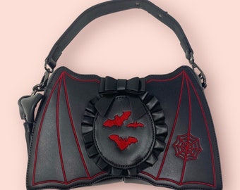 May include: Black faux leather handbag with red stitching, shaped like bat wings with a red bow and bat embroidery.