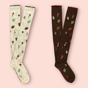 May include: Two pairs of knee-high socks with a mushroom and leaf pattern. One pair is cream with brown mushrooms and leaves, and the other pair is brown with cream mushrooms and leaves.