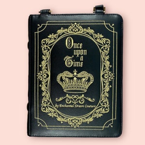 May include: Black faux leather book-shaped crossbody bag with gold embroidery. The embroidery features a crown, ornate scrollwork, and the text "Once upon a Time by Enchanted Dream Couture".