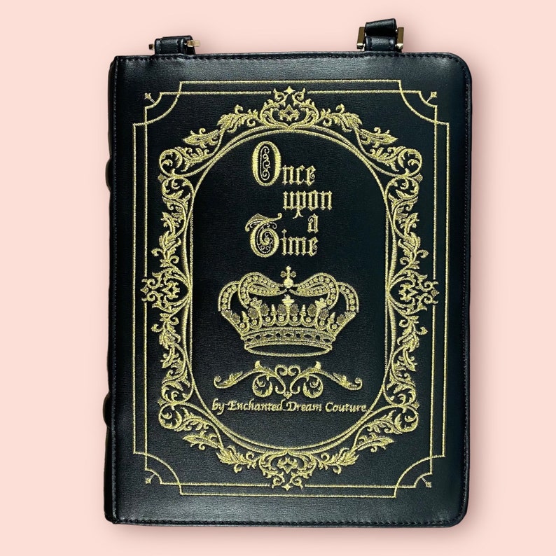 May include: Black faux leather book-shaped crossbody bag with gold embroidery. The embroidery features a crown, ornate scrollwork, and the text "Once upon a Time by Enchanted Dream Couture".