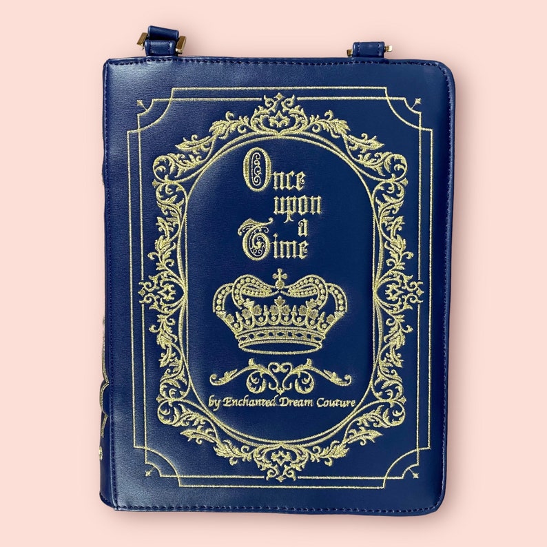 May include: A blue faux leather book clutch with a gold embroidered design. The design features a crown, ornate scrollwork, and the text "Once upon a Time" by Enchanted Dream Couture.