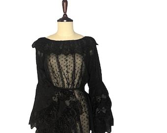 Puede incluir: Top de encaje negro con mangas largas y acampanadas y un corpiño transparente. El top presenta intrincados detalles de encaje alrededor del escote, la cintura y las mangas, con pequeños lazos adornando las mangas. La prenda se exhibe en un maniquí.