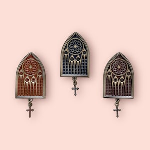 Peut inclure: Trois épingles en émail doré en forme de vitraux avec un charme de croix suspendu à chacune. Les fenêtres sont dans des tons de rouge, bleu et marron.
