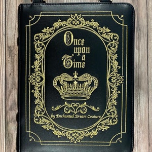 May include: Black leather book-shaped bag with a gold embroidered design. The design features a crown, ornate scrollwork, and the text "Once upon a Time" by Enchanted Dream Couture.