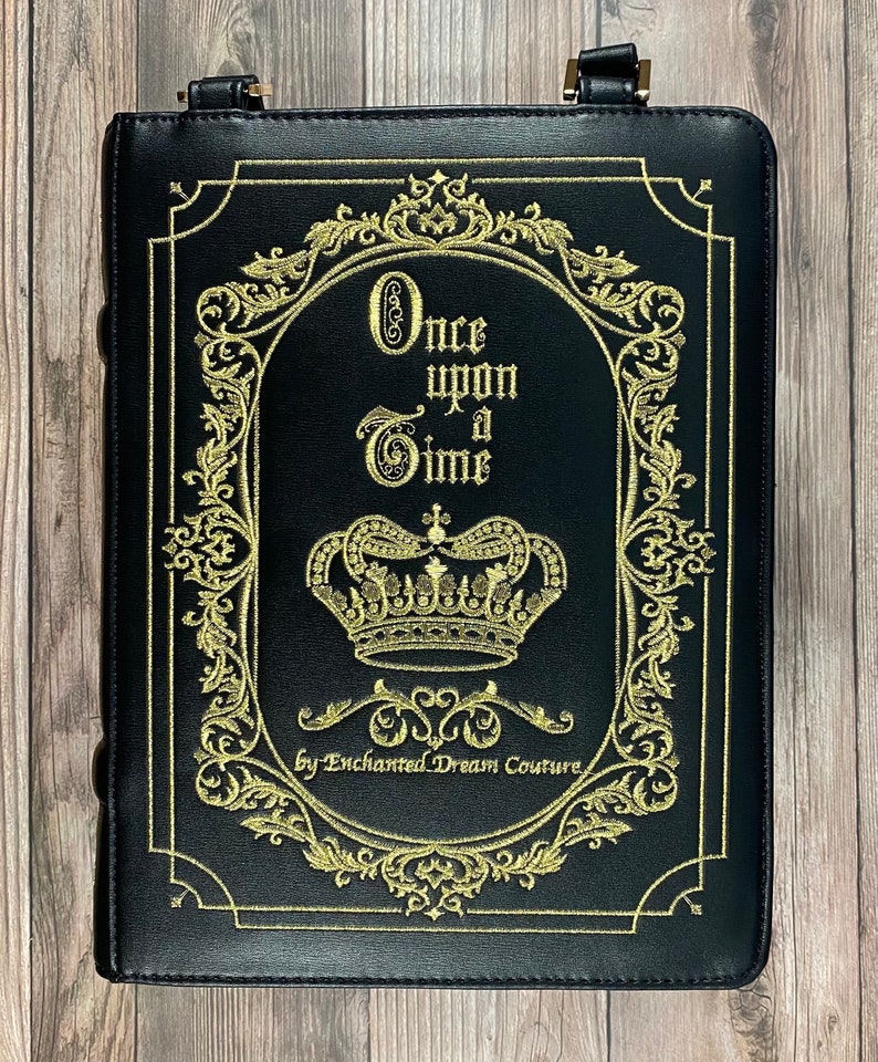 May include: Black leather book-shaped bag with a gold embroidered design. The design features a crown, ornate scrollwork, and the text "Once upon a Time" by Enchanted Dream Couture.