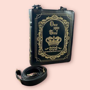 May include: Black faux leather book-shaped bag with a gold chain strap and gold embroidery that reads "Once upon a Time" with a crown and "By Enchanted Dream Couture".