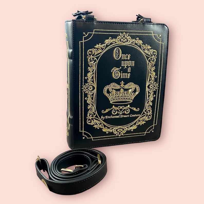 May include: Black faux leather book-shaped bag with a gold chain strap and gold embroidery that reads "Once upon a Time" with a crown and "By Enchanted Dream Couture".