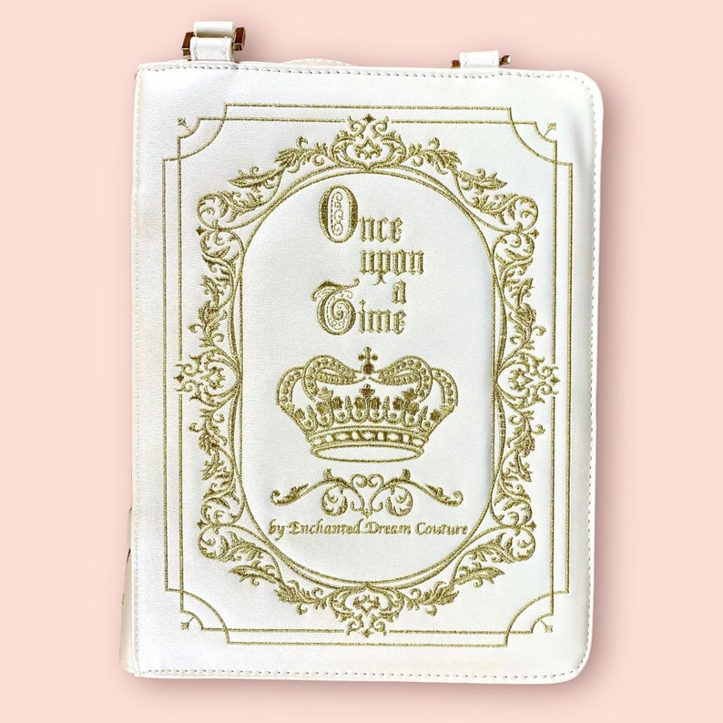 May include: White faux leather book clutch with a gold embroidered design. The design features a crown, ornate scrollwork, and the text "Once upon a Time" by Enchanted Dream Couture.