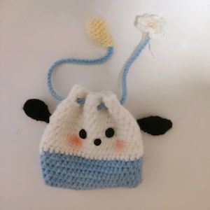 Könnte beinhalten: Eine kleine gehäkelte Kordeltasche mit einem weißen und blauen Design. Die Tasche hat schwarze Ohren und ein weißes Gesicht mit schwarzen Augen und einer schwarzen Nase. Die Tasche hat einen hellblauen Riemen mit einer gelben Blume am Ende.