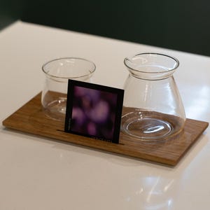 May include: A wooden serving tray with two clear glass vessels and a small card. One vessel is a small, round glass, and the other is a carafe with a spout. The card has a blurred purple image and the text "DOLCE VITA".