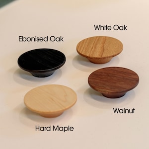 Puede incluir: Cuatro pomos de madera para armarios en diferentes tipos de madera: Roble ebanizado, Roble blanco, Nogal y Arce duro. Cada pomo es redondo con una base cilíndrica corta. Los pomos se muestran sobre una superficie clara.