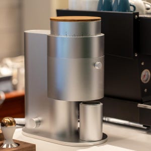 Peut inclure: Un moulin à café argenté avec un dessus en bois et un design cylindrique. Le moulin a un cadran avec des marquages de mesure et un petit récipient argenté à la base. Une machine à expresso noire est en arrière-plan.