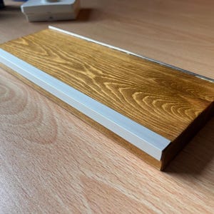 May include: A rectangular wooden shelf with a warm brown wood grain finish, featuring a thin, white, L-shaped metal trim along the front edge. The shelf has a smooth, polished surface and a clean, linear design with a flat top and sides.