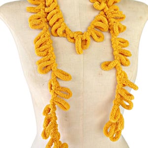 May include: A long, yellow, crocheted scarf with a textured, fuzzy look. The scarf is draped around a mannequin's neck and hangs down to the waist.