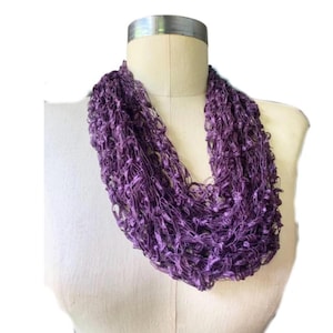 May include: A purple infinity scarf made of a loosely woven, textured material. The scarf is displayed on a mannequin and has a looped design. The color is a deep, rich shade of purple, suitable for adding a pop of color to any outfit.