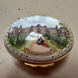 May include: An oval-shaped trinket box with a gold-colored rim. The lid features a detailed illustration of Vassar College, with a horse-drawn carriage on a path leading to the building. The text "VASSAR COLLEGE" is displayed on a banner.
