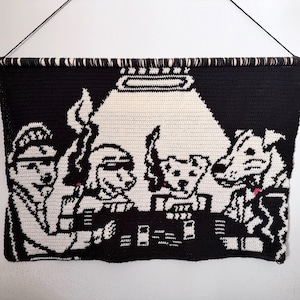 May include: A black and white crochet wall hanging showing dogs playing poker. The artwork is mounted on a wooden dowel and suspended by a black cord. The dogs are in a poker game, with cards and chips on the table.