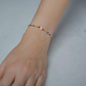 Könnte beinhalten: Ein Armband mit Perlen in Regenbogenfarben. Das Armband besteht aus kleinen, bunten Perlen mit einem weißen Blumenanhänger in der Mitte. Das Armband wird am Handgelenk getragen.