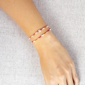 Könnte beinhalten: Zwei Armbänder aus Perlen in Rosa, Orange und Weiß werden am Handgelenk getragen. Ein Armband hat einen kleinen Blumenanhänger. Der Hintergrund ist hellgrau.
