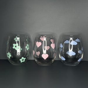 Op de afbeelding: Drie wijnglazen zonder voet met decoratieve ontwerpen. Eén glas heeft lichtgroene bloemen, een ander roze harten en de derde blauwe wolken. De glazen zijn helder en staan op een donkere ondergrond.