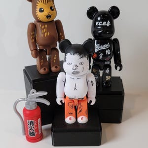 May include: Three black, white, and orange bear figurines with black eyes and black ears. The bear on the left is brown with a white ear. The bear in the middle has a white body with a black face and orange pants. The bear on the right is black with white text that says "F.C.R.B. Bristol". The bears are sitting on black square bases. A red fire extinguisher with Japanese text is in the foreground.