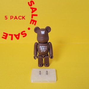 May include: A brown bear-shaped toy with a white "W" on its chest and Japanese text on its shirt. The toy is standing on a white display stand with two silver pins.