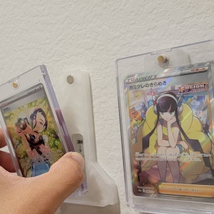 May include: Three Pokemon trading cards are displayed in clear plastic protective cases mounted on a white wall. The cards are all different and feature different characters. The card on the left is a close-up of a female character with a pink and blue outfit. The card in the center features a female character with long black hair and a yellow outfit. The card on the right features a character with a red and orange outfit. The text on the center card reads "Trainer's - カミツレのきらめき - Fusion Arts".