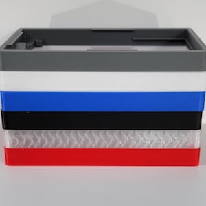 May include: A stack of six different colored plastic boxes. The boxes are gray, white, blue, black, white with a textured pattern, and red.