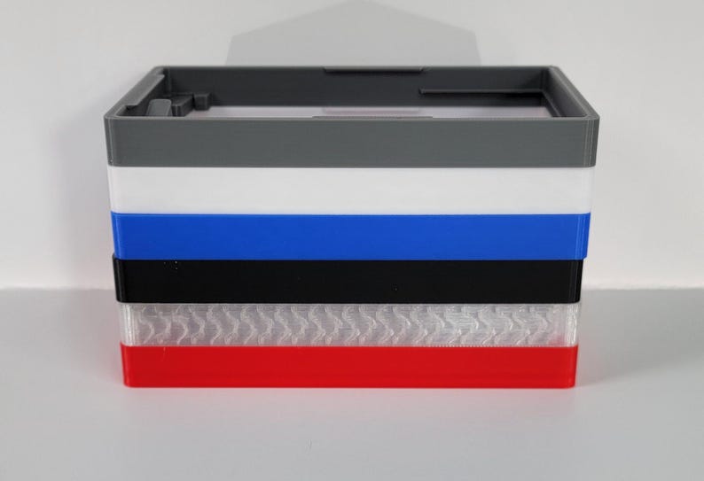 May include: A stack of six different colored plastic boxes. The boxes are gray, white, blue, black, white with a textured pattern, and red.