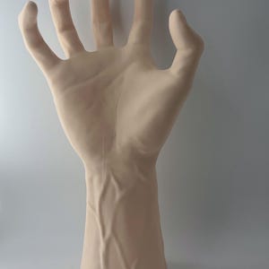 May include: A realistic-looking, pale-toned, sculpted hand with veins, posed with fingers extended and slightly splayed. The hand is attached to a forearm, and the sculpture is made of a smooth, matte material.