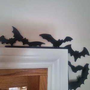 May include: Black bat decorations on a white door frame and wall. The bats are arranged in a decorative pattern, ideal for Halloween. The bats are made of a dark material and are silhouetted against the white background.