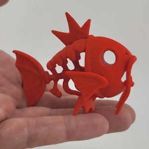 May include: A 3D-printed, bright red fish figurine with a skeletal design. The fish has a crown-like structure on its head, fins, and a tail. The figurine is held in a hand, showcasing its details and form.