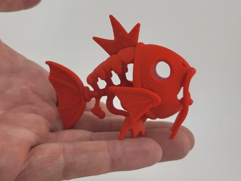 May include: A 3D-printed, bright red fish figurine with a skeletal design. The fish has a crown-like structure on its head, fins, and a tail. The figurine is held in a hand, showcasing its details and form.