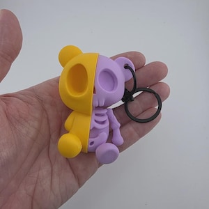 Peut inclure: Porte-clés en forme d'ours bicolore, jaune et violet. La moitié de l'ours est un squelette. Le porte-clés est fixé à un anneau en métal noir. Objet décoratif.