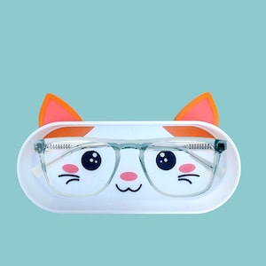 Könnte beinhalten: Eine Brille mit transparentem Rahmen liegt in einem weißen, katzenförmigen Etui mit orangefarbenen Ohren. Das Etui hat ein Cartoon-Katzen-Gesicht mit schwarzen Augen, rosa Wangen und einem kleinen Mund. Der Hintergrund ist hellblau.