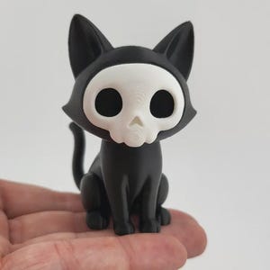 Op de afbeelding: Een zwart kattenbeeldje met een wit schedelgezicht. De kat zit rechtop met zijn staart gekruld. Het beeldje is ongeveer 10 cm hoog en is gemaakt van een stevig materiaal.