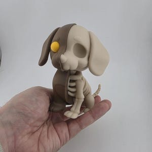 Peut inclure: Figurine originale d'un chien avec un design squelette. Le chien est divisé en deux, une moitié marron et l'autre beige, avec un œil jaune. Le squelette est visible sur le côté beige. La figurine est tenue dans une main sur fond blanc.
