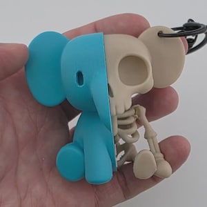 Peut inclure: Un porte-clés original avec un éléphant bleu d'un côté et un squelette de l'autre. La forme de l'éléphant est divisée, révélant la structure squelettique. Le porte-clés est maintenu par un anneau noir.