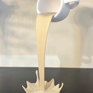 May include: A white ceramic cup pouring a stream of beige liquid into a beige ceramic splash. The cup is tilted upside down and the liquid is frozen in mid-air.