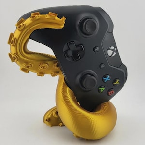 May include: A black Xbox controller rests on a gold-colored octopus-shaped stand. The stand has a detailed, tentacle design. The controller has colored buttons and a directional pad.