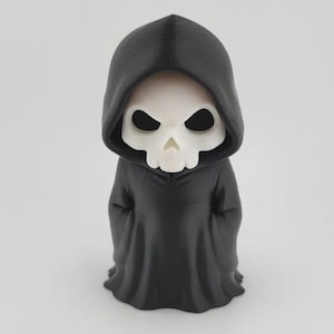 Op de afbeelding: Een beeldje van de Grim Reaper, met een wit schedelgezicht en zwarte oogkassen, gekleed in een zwarte capuchon. De figuur staat rechtop, met de kap over het hoofd en de schouders. Het ontwerp is een gestileerde, cartoonachtige weergave.