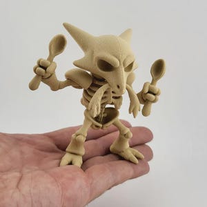 Peut inclure: Figurine beige d'une créature squelettique tenant deux cuillères. La figurine a une tête en forme de crâne avec des oreilles pointues et une cage thoracique. La figurine est tenue dans la main d'une personne.