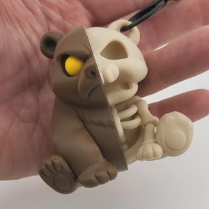 Peut inclure: Pendentif avec un design divisé : un côté est un ours brun, l'autre un squelette blanc os. L'ours a un œil jaune. Le pendentif est suspendu à un cordon noir, présentant un mélange unique de mignon et de macabre.