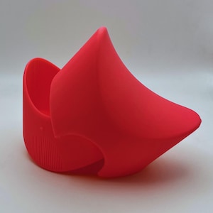 K&ouml;nnte beinhalten: Ein leuchtend roter Topfuntersetzer aus Silikon mit einem einzigartigen, geschwungenen Design. Der Halter hat eine abgerundete Basis mit vertikalen Rippen und eine spitze, abstrakte Form, die sich nach oben erstreckt. Die Gesamtform ist glatt und modern.