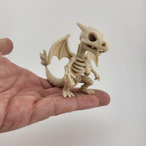 Könnte beinhalten: Beige Skelett-Drachenfigur mit detailliertem Brustkorb, Flügeln und Schwanz. Der Drache befindet sich in einer hockenden Position. Die Figur wird in einer Hand vor einem schlichten Hintergrund gehalten. Der Drache ist ein Fantasy-Themenartikel.
