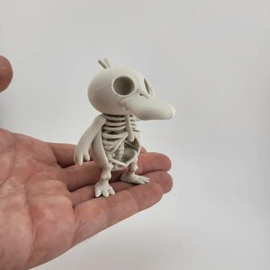 May include: A small, off-white figurine of a duck skeleton. The duck has a long beak, round eyes, and a detailed rib cage. The figurine is held in a person's hand against a plain white background. The duck is a whimsical, Halloween-themed decoration.